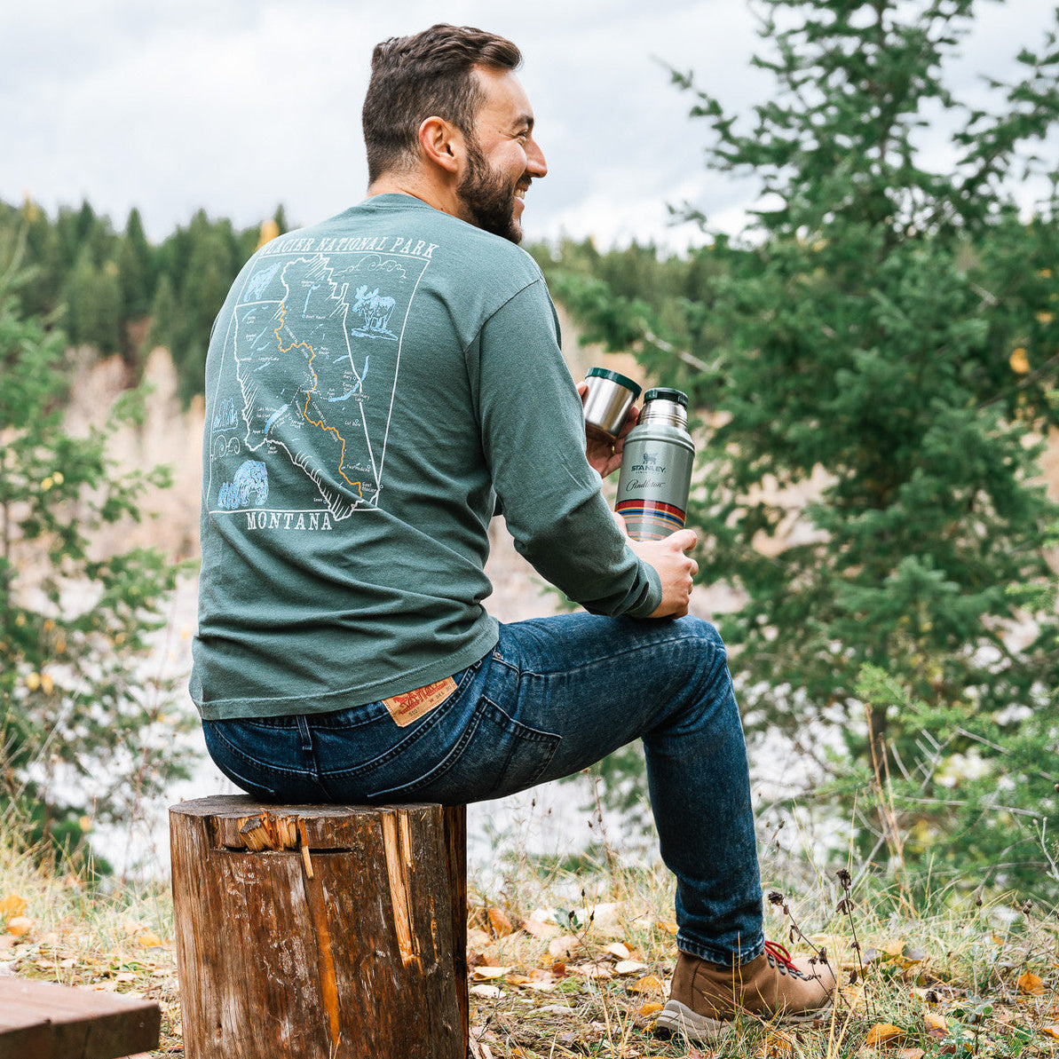Man sitting on a log in a forest, wearing a green t-shirt with a map design, holding a thermos.